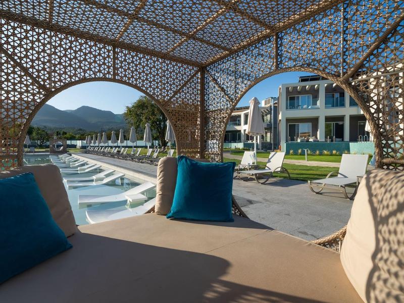 Pohled z krytého odpočinkového prostoru na hotelový bazén s lehátky a budovami v pozadí.