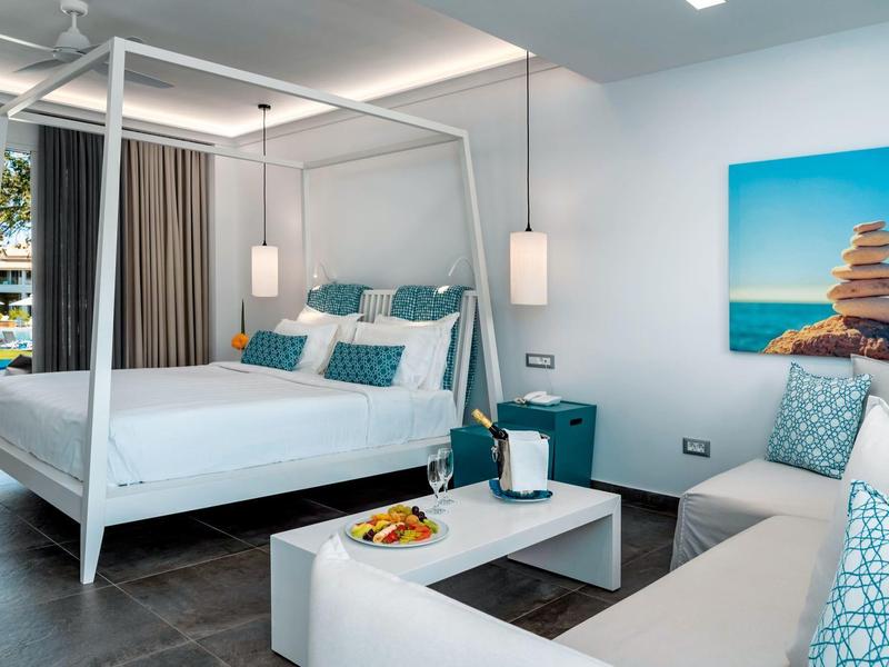 Moderně zařízený hotelový pokoj s nebeskou postelí, pohovkou a stolem s ovocem.