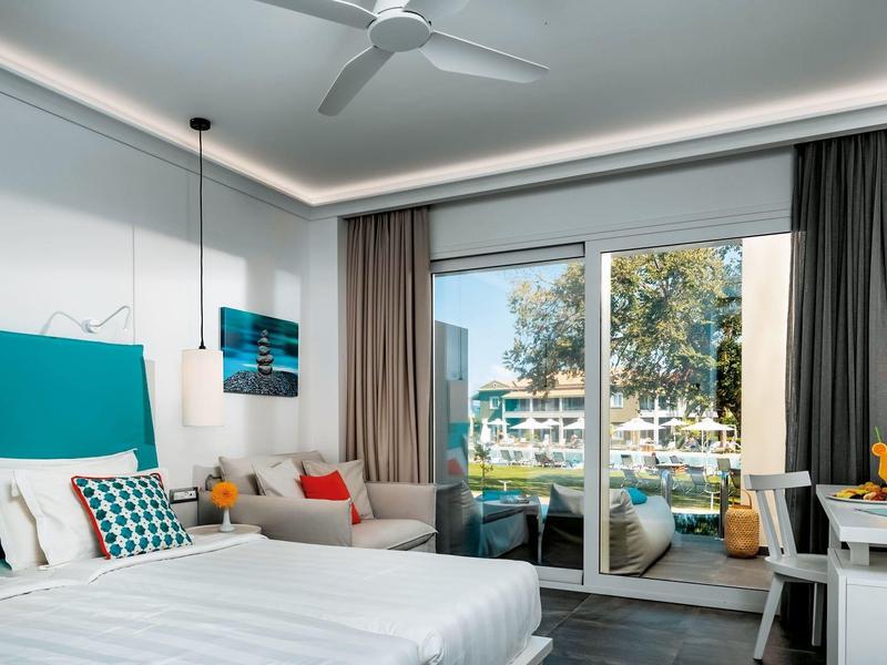 Hotelový pokoj se dvěma postelemi, velkým balkónem a výhledem na bazén a stromy.