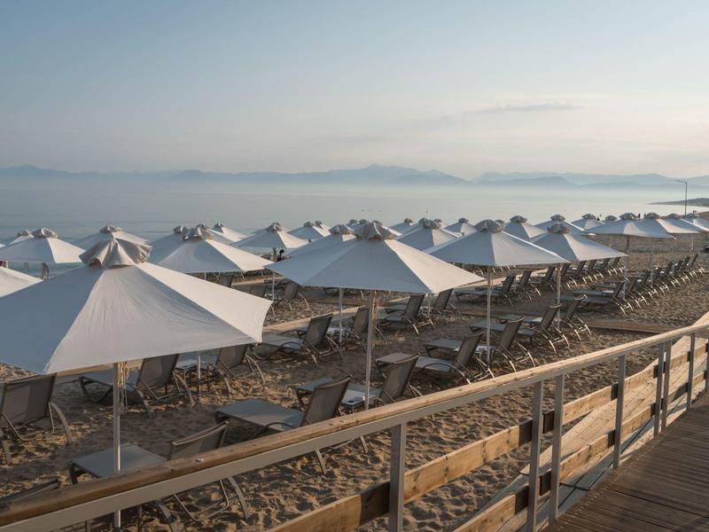 Řady bílých slunečníků na pláži s mořem a horami v pozadí.