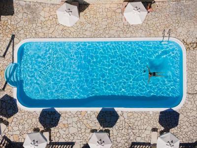 Piscine d'hôtel ovale avec eau bleue claire, entourée de parasols sur un sol en mosaïque de pierre.