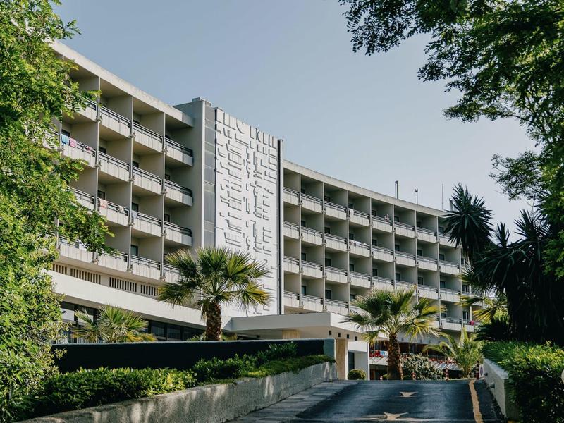 Mehrstöckiges Hotelgebäude mit Balkonen, Palmen und üppigem Grün an sonnigem Tag.