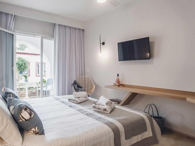 Moderní hotelový pokoj s manželskou postelí, televizí na stěně a přístupem na balkon s rostlinami.