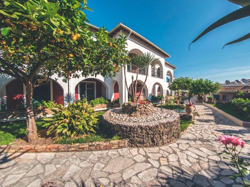 Dvoupodlažní bílá hotelová budova s ​​zahradou, kamennou cestou a fontánou pod jasnou oblohou.