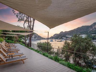 Terraza con tumbonas y velas de sombra con vistas a una bahía y montañas al atardecer.