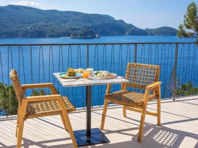 Terras met twee houten stoelen en een tafel met eten, uitzicht op blauwe zee en bergen.