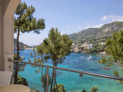 Vista de una bahía azul con barcos desde un balcón con barandilla de vidrio y mesas.