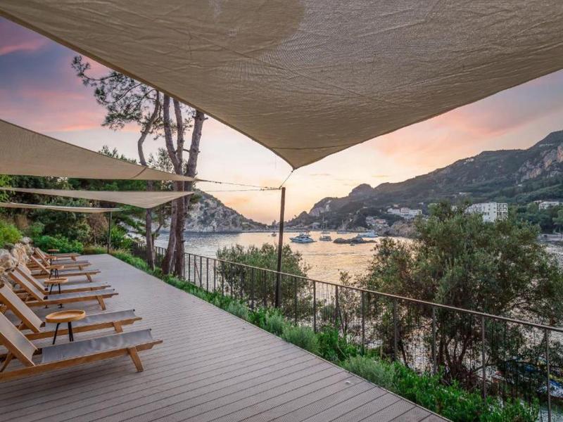 Terras met ligstoelen met uitzicht op een baai bij zonsondergang met zeilboten en heuvels.