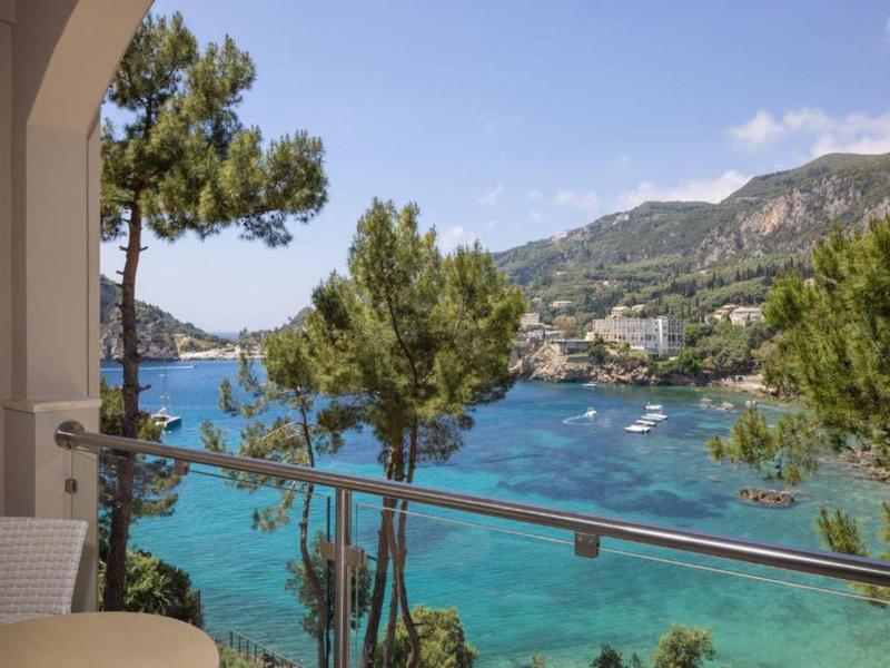 Balkonzicht op de zee met boten en groene heuvels onder een blauwe lucht.