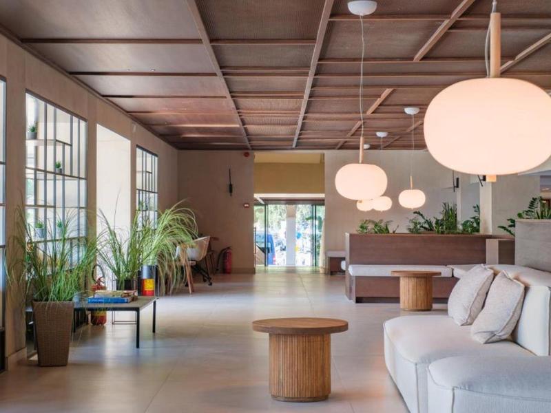 Lichte hotel lobby met grote ramen, planten, houten plafond, witte bank en ronde hanglampen.