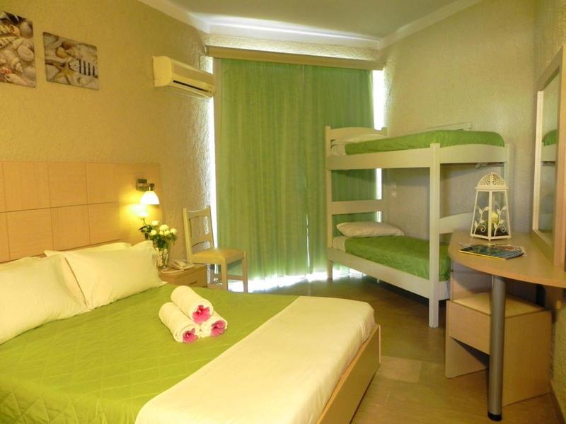 Hotelový pokoj s manželskou postelí, palandou, psacím stolem a zelenými závěsy.