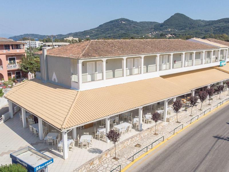 Moderní hotelová budova s terasou a markýzami podél silnice v slunné krajině.