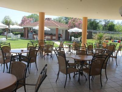 Covered terrace with tables and chairs, green garden and buildings in the background.