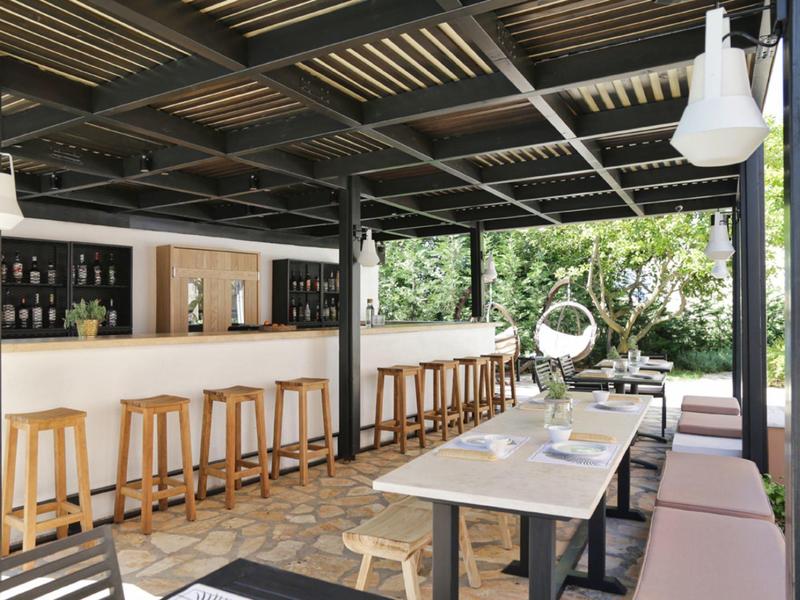 Nowoczesna przestrzeń na zewnątrz z barem, drewnianymi stołkami i stołami w zadaszonym miejscu z widokiem na zieleń.