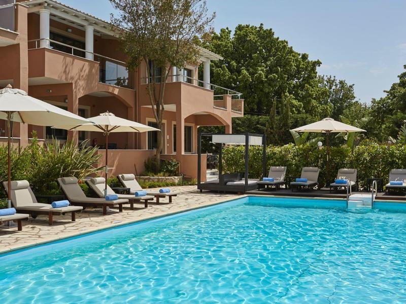 Basen hotelowy z leżakami i parasolami obok wielopiętrowego budynku i drzew.