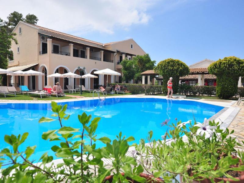 Hotel met groot buitenzwembad, parasols en planten onder een zonnige hemel.