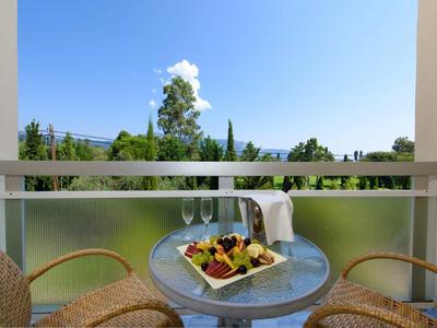 Balkon mit kleinem Glas-Tisch, zwei Stühlen und Teller mit Obst, Blick auf grüne Bäume und blauen Himmel.