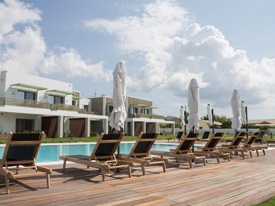 Ein moderner Poolbereich mit Sonnenliegen und geschlossenen Sonnenschirmen vor einem Hotelgebäude unter einem bewölkten Himmel.