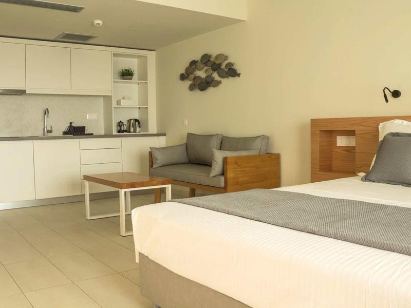 Modernes, helles Zimmer mit Küchenzeile, grauem Sofa, Holzmöbeln und großem Bett mit grauem Bettläufer.