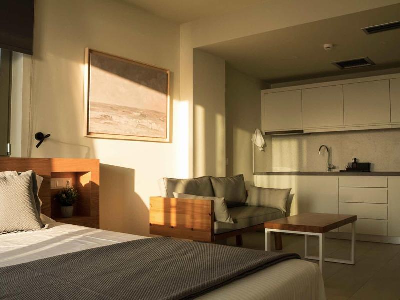 Ein modernes Schlafzimmer mit großem Bett, Sofa, Tisch und heller Küchenzeile im Hintergrund.