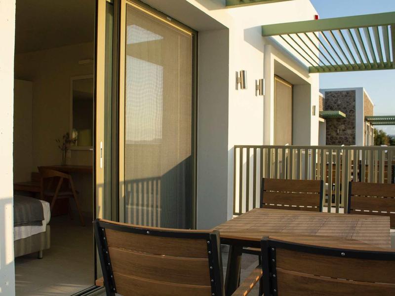 Balkon mit Holztisch und Stühlen, Gebäude mit großen Fenstern, Sonnenschutz aus Metall.