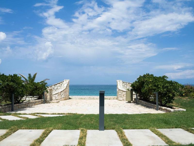 Strandzugang mit weißem Sand, grünem Gras, blauen Himmel und Meer im Hintergrund.