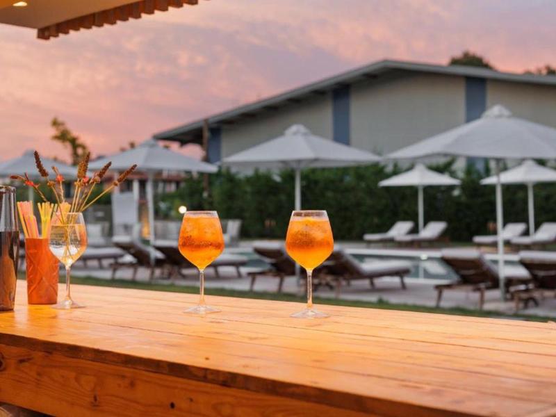 Drei Orangencocktails stehen auf einer Holztheke vor Liegestühlen und Sonnenschirmen bei Sonnenuntergang.