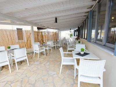 Zastřešená terasa s bílými židlemi a stoly vedle budovy s velkými okny.