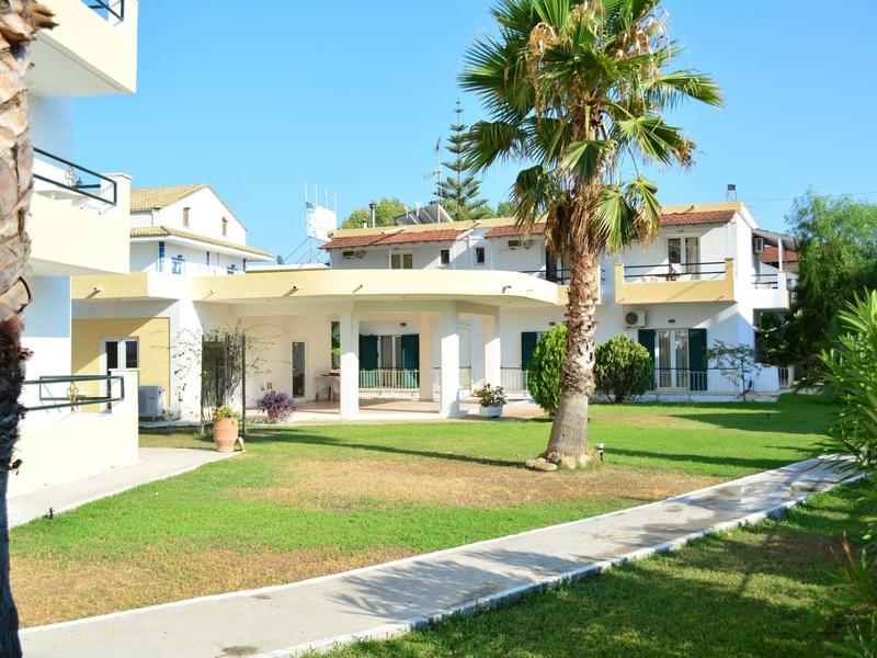 Moderní rekreační komplex s bílými byty, palmami a upravenými trávníky pod jasnou oblohou.