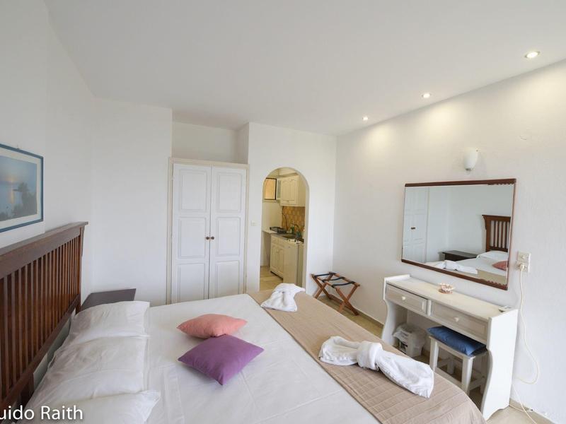 Helles Schlafzimmer mit Doppelbett, Wandspiegel, Kommode, Bademänteln und Tür zum Bad.