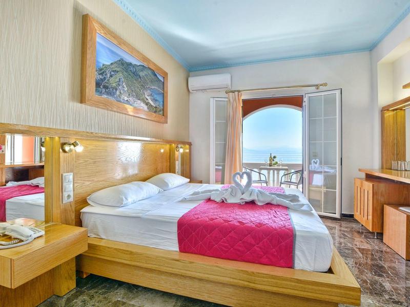 Chambre d'hôtel lumineuse avec lit double, couvre-lit rouge, balcon et vue sur la mer.