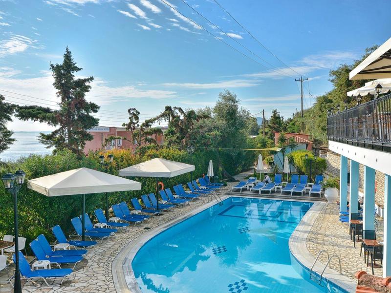 Piscine extérieure avec chaises longues bleues et parasols entourée d'arbres et de bâtiments sous un ciel dégagé.