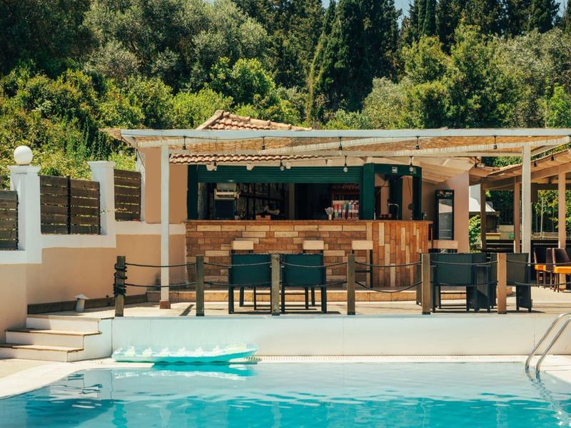Piscine extérieure avec bar et chaises entourée d'arbres verts et ensoleillement.