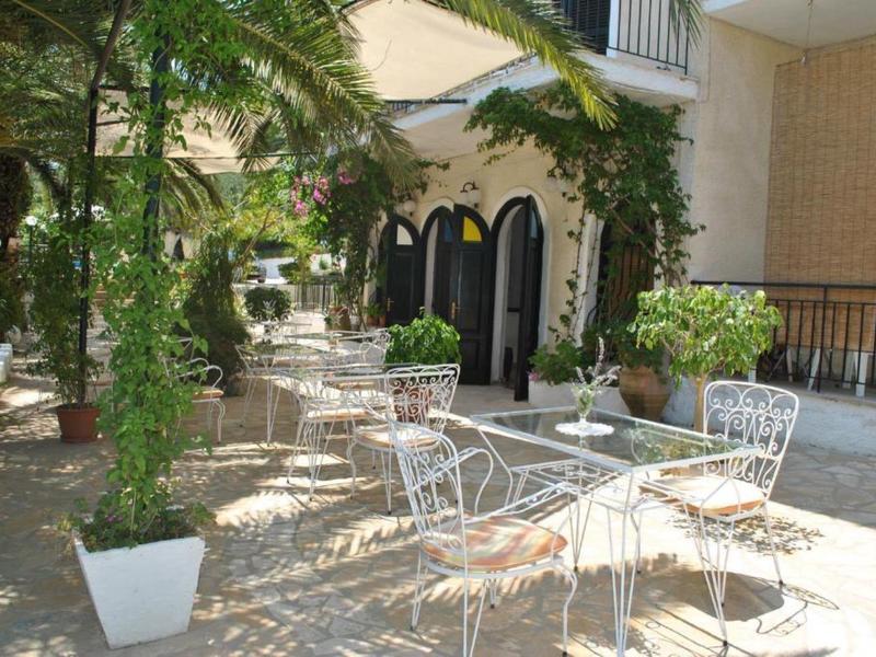 Slunná terasa s bílými kovovými stoly a židlemi, obklopená rostlinami.