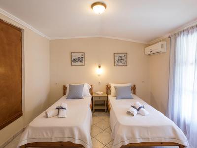 Zwei Einzelbetten mit weißen Decken, Holzmöbel, Vorhang an Fenster, Wandbilder, Klimaanlage.