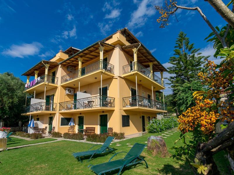 Drie verdiepingen tellend hotel met balkons en ligstoelen in de tuin onder een blauwe lucht.
