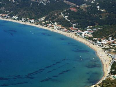 Letecký pohled na zakřivenou písečnou pláž s čistou modrou vodou a okolními budovami