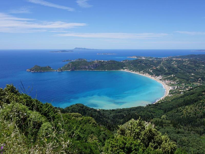 Pohled na zakřivené pobřeží s modrou vodou a hustě zalesněnými kopci pod modrou oblohou.