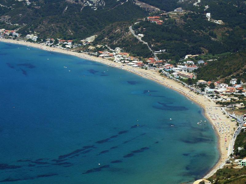 Letecký pohled na zakřivenou písečnou pláž s čistou modrou vodou a okolními budovami