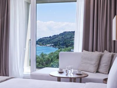 Bright hotel room with view of sea and green hills through large windows.