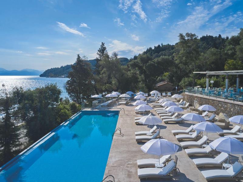 Pool with white loungers and umbrellas by the sea in a wooded setting.