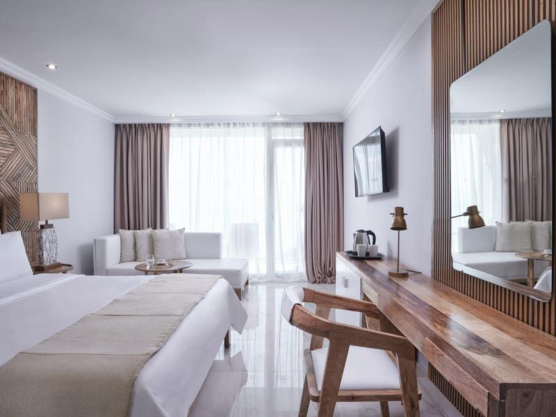 Bright hotel room with double bed, desk, chair, and large window with curtains.