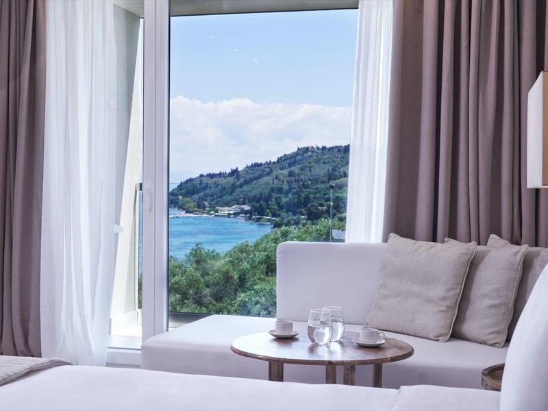 Bright hotel room with view of sea and green hills through large windows.
