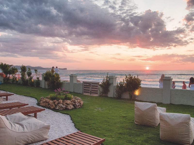 Vue sur la plage au coucher du soleil avec chaises longues et poufs sur pelouse.