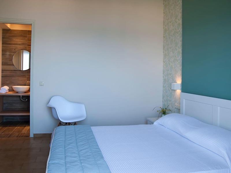 Chambre d'hôtel moderne avec grand lit, chaise blanche et vue sur une salle de bain adjacente.
