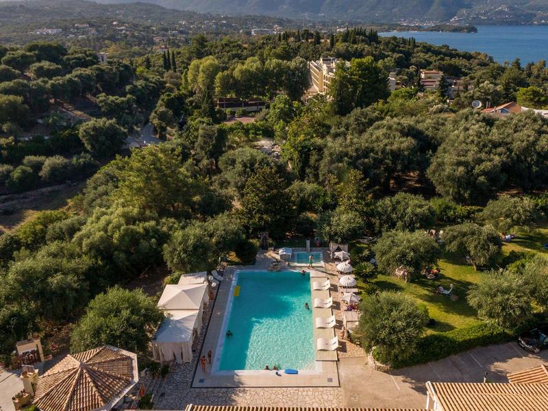 Aerial view of a hotel with a large pool, surrounded by trees and overlooking the sea.