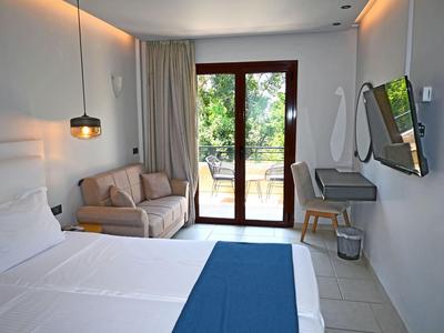 Helde hotelkamer met tweepersoonsbed, bank, glazen deur naar tuin en eenvoudige inrichting.