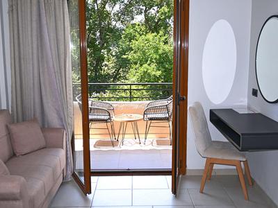 Lichte hotelkamer met balkon, bank, stoelen en uitzicht op groene bomen.