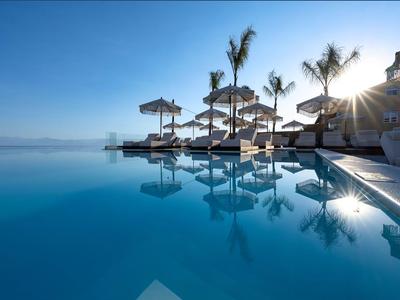 Infinity zwembad met ligbedden en rieten parasols onder een heldere lucht bij een hotel.
