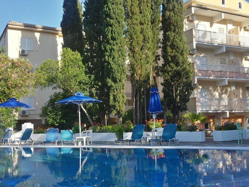 Edificio del hotel con balcones rodeado de cipreses y piscina con tumbonas y sombrillas.